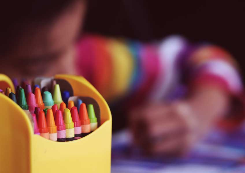 A box of crayons in front of a child who is coloring